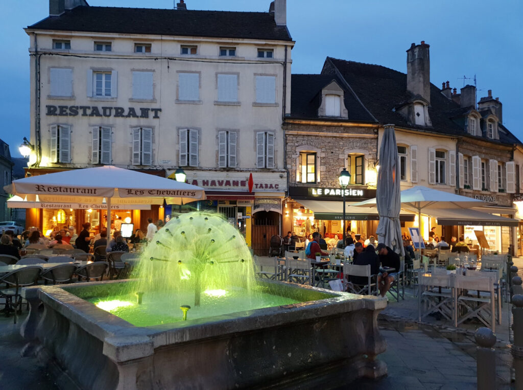 Avondfoto van een verlicht plein in Beaune met fonteinbol, historische Bourgondische panden en sfeervolle terrassen met dinerende gasten.