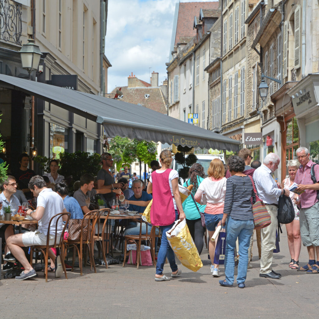 Zonnige dagfoto van het oude centrum van Beaune met winkelende mensen, terrassen en levendige sfeer.