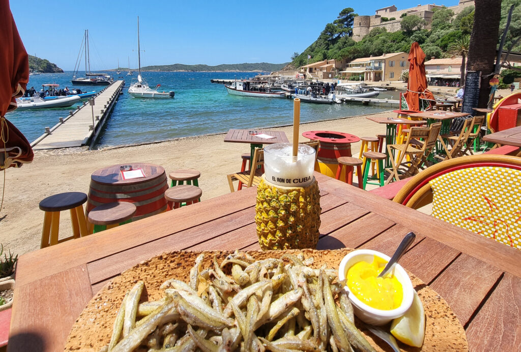 Platte mit frittierten Sardellen und Tapenade, eine Pina Colada in einer Ananas, Barhocker, schwimmender Holzsteg mit Segelbooten, U-Boot mit Tauchern, Fort du Moulin und das Hafenmeistergebäude auf Port-Cros.
