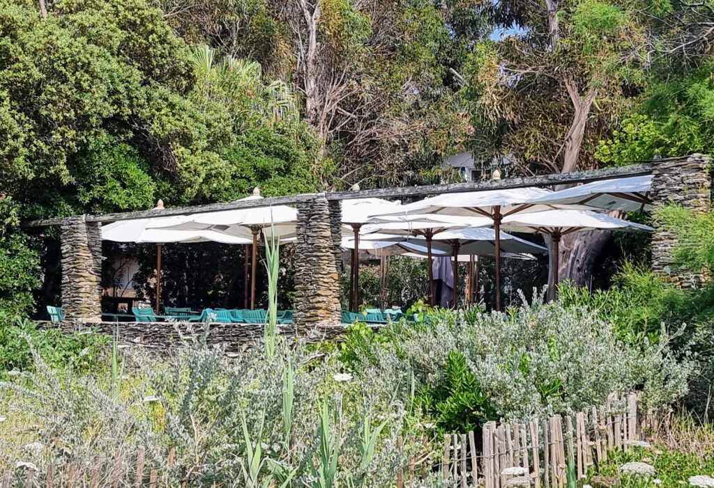 Stimmungsfoto der Terrasse des Hotels Le Manoir auf Port-Cros, gesehen von Gras und Büschen aus, mit gestapelten Steinsäulen, grünen Stühlen und weißen Sonnenschirmen.