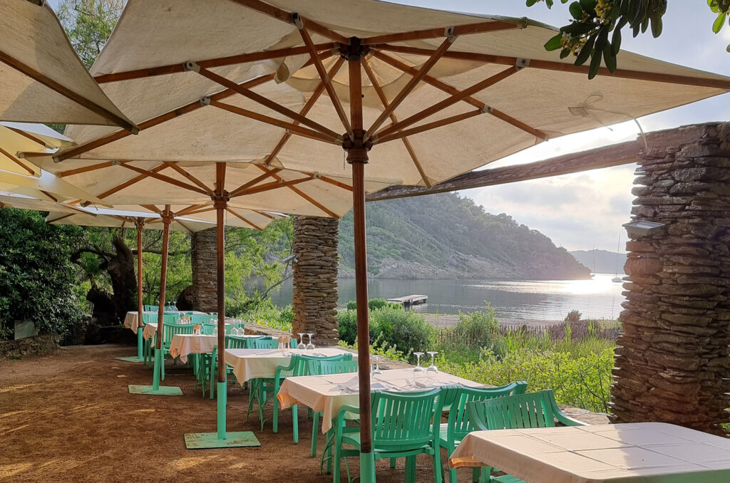 Restaurant extérieur du Manoir à Port-Cros avec des piliers en pierre, de grands parasols, des tables recouvertes de nappes blanches et une vue sur la baie, un ponton en bois, la mer et les collines boisées.