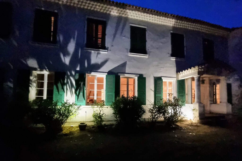 Photo nocturne de la façade de l'hôtel Le Manoir à Port-Cros, avec sa façade illuminée, ses volets verts et son entrée à colonnes.
