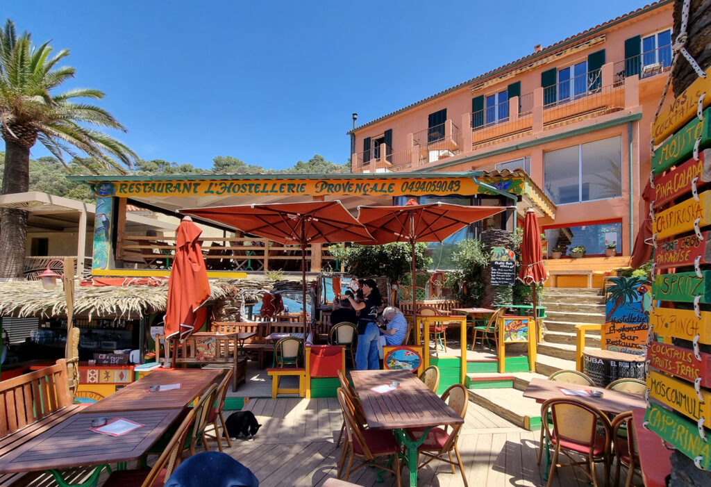 Terrasse der Hostellerie Provençale auf Port-Cros mit Holztischen, bunten Stühlen, einem Kellner, der bedient, und einem fröhlichen Cocktailschild im Hintergrund.