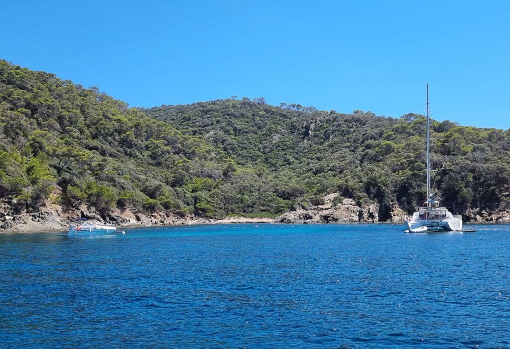 Blick vom Meer auf die zerklüftete Küste von Port-Cros mit Felsen, bewaldeten Hängen, einem Katamaran rechts, einem kleinen Sportboot links und der abgesperrten Zone zum Unterwassermuseum.