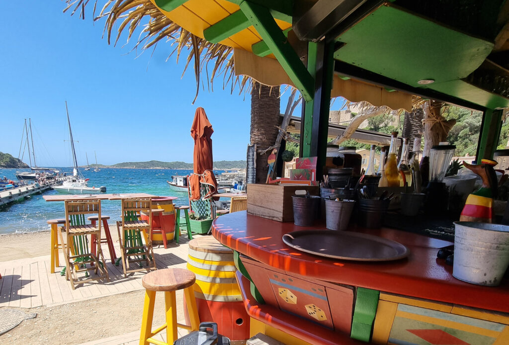 Bar coloré de l'Hostellerie Provençale dans le port de Port-Cros, avec tabourets de bar, tables, petite promenade, mer bleu azur et vue sur Hyères.