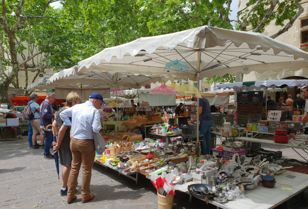 Markt in Uzès met kraampjes onder grote parasols, platanen en mensen die naar de uitgestalde producten kijken.