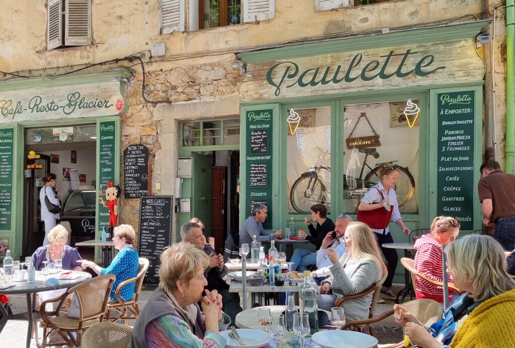 Gezellig terras in het centrum van Cotignac waar mensen eten onder de schaduwrijke platanen, met een restaurant op de achtergrond