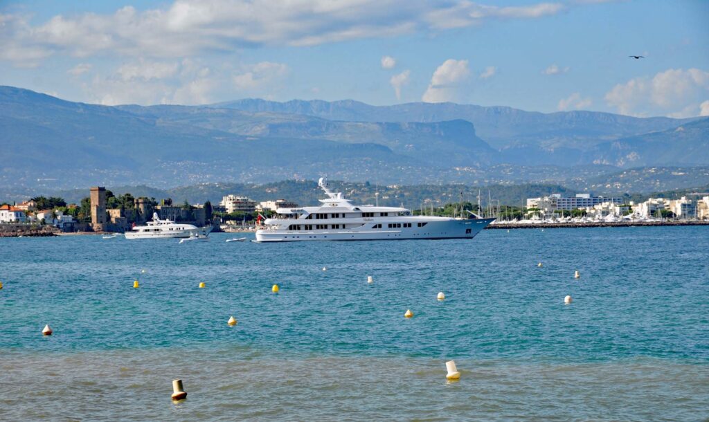 Un yacht élégant flotte dans les eaux bleu azur de la baie de La Napoule, offrant une vue imprenable sur le littoral. Luxurious yacht anchored in the bay of La Napoule with clear blue water and coastline in the background.