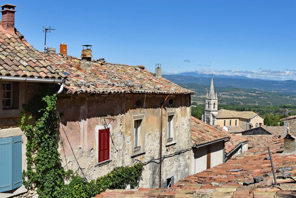 View of Bonnieux with historic buildings, a narrow street sloping downwards and Mont Ventoux in the distance.