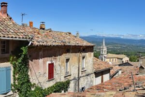 View of Bonnieux with historic buildings, a narrow street sloping downwards and Mont Ventoux in the distance.