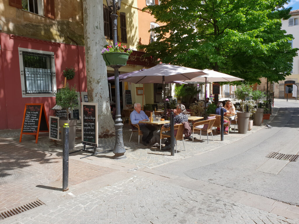 De hoofdstraat van het Provençaalse dorp Lorgues met historische panden, terrasjes met parasols en mensen die buiten zitten.