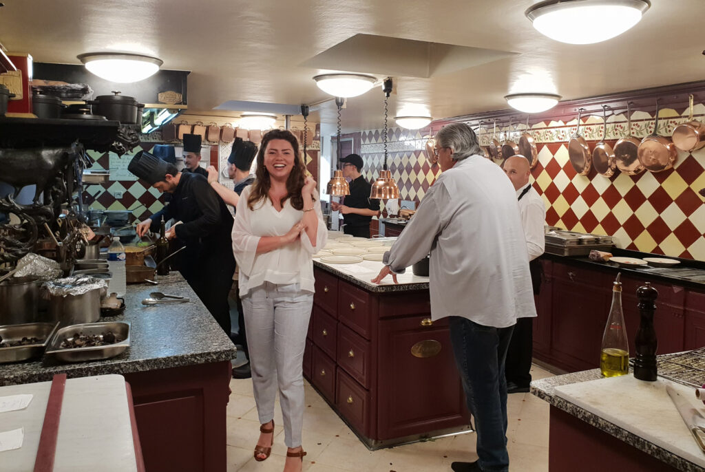 Jonge vrouw die blij poseert in de keuken van Restaurant Bruno, met chefs die op de achtergrond aan het werk zijn