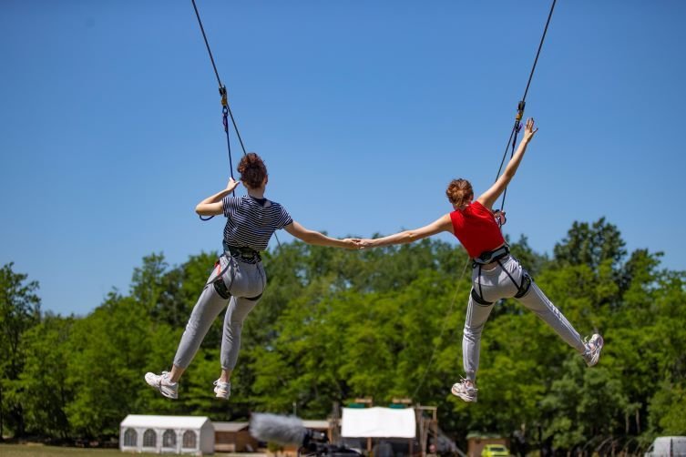 Twee meisjes die tegelijk van een zipline afglijden in Accroche Aventure, terwijl ze elkaars hand vasthouden.