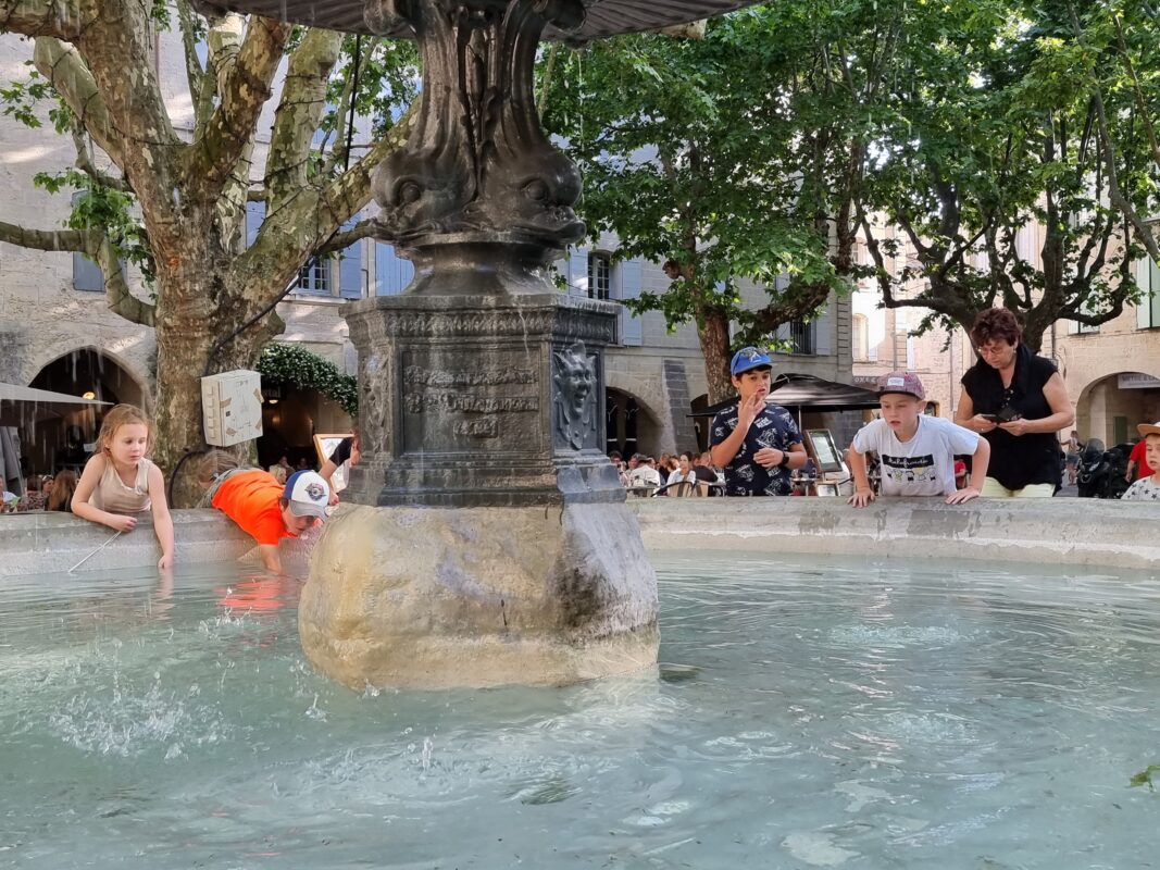 Des enfants accrochés au muret de la fontaine de la place aux Herbes à Uzès jouent avec l'eau, avec en arrière-plan des terrasses et des platanes.