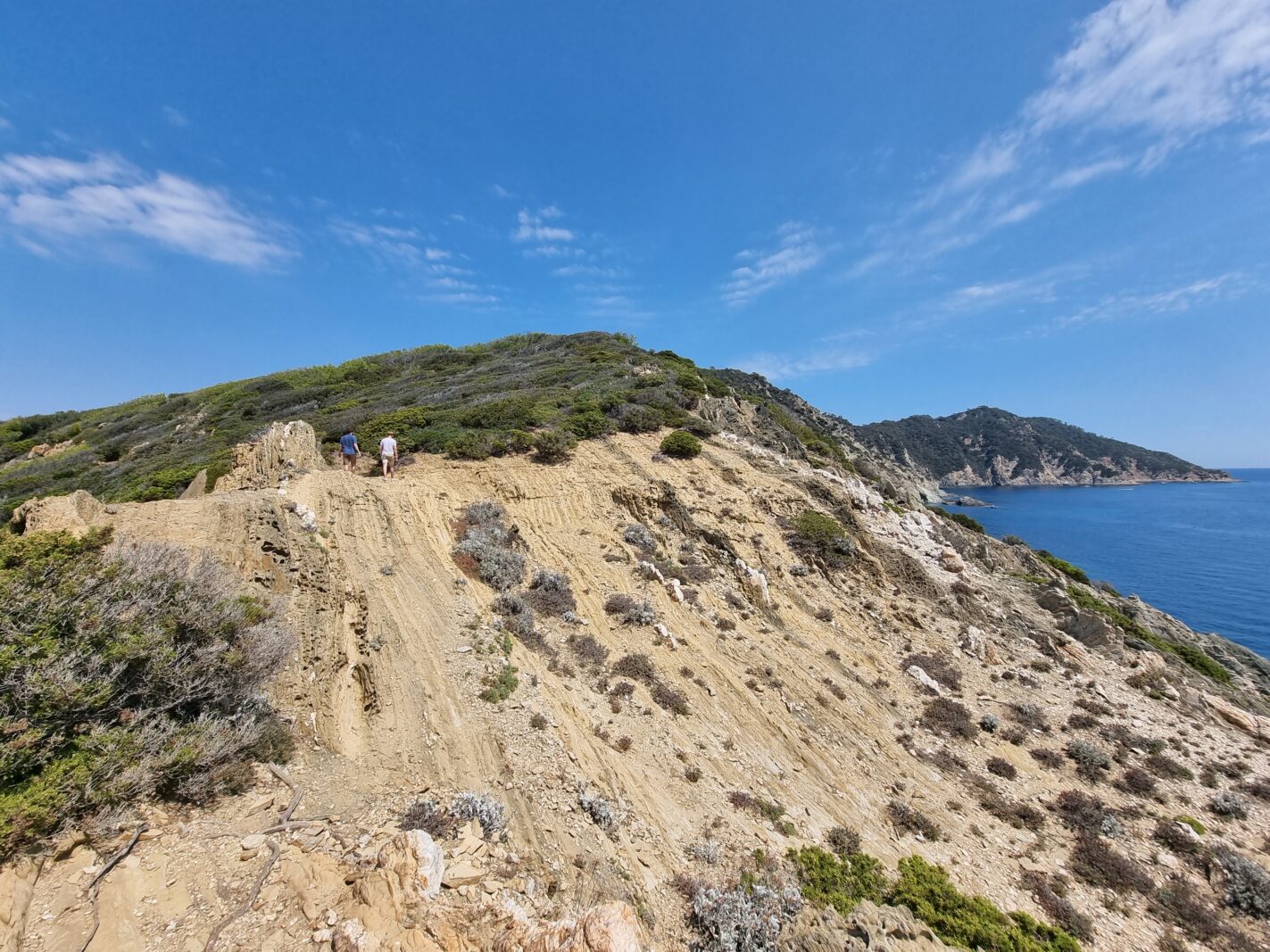 Wandeling op Port-Cros met uitzicht over een kloof, rotswand aan de overzijde, twee wandelaars op de rug en begroeiing met de zee rechts in beeld.